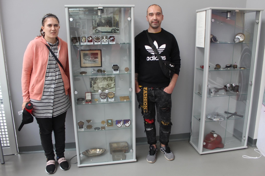 Uczestniczka wraz z Uczestnikiem pozują do zdjęcia na tle ekspozycji z częściami samochodowymi. 