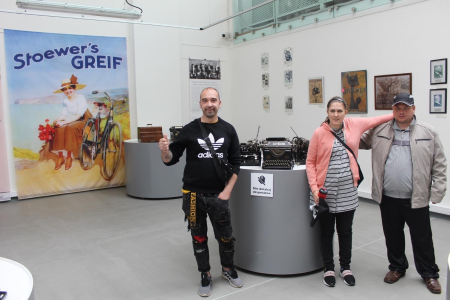 Uczestniczka wraz z Uczestnikami pozuje do zdjęcia na tle wystawy z maszynami do pisania.