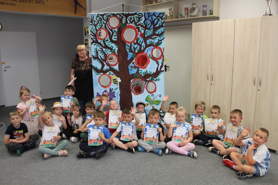 Grupa dzieci wraz z opiekunem trzymająca dyplomy pozuje do zdjęcia siedząc.