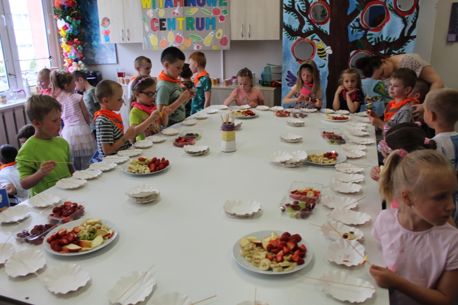 Grupa dzieci jedząca owocowe szaszłyki przy stole.