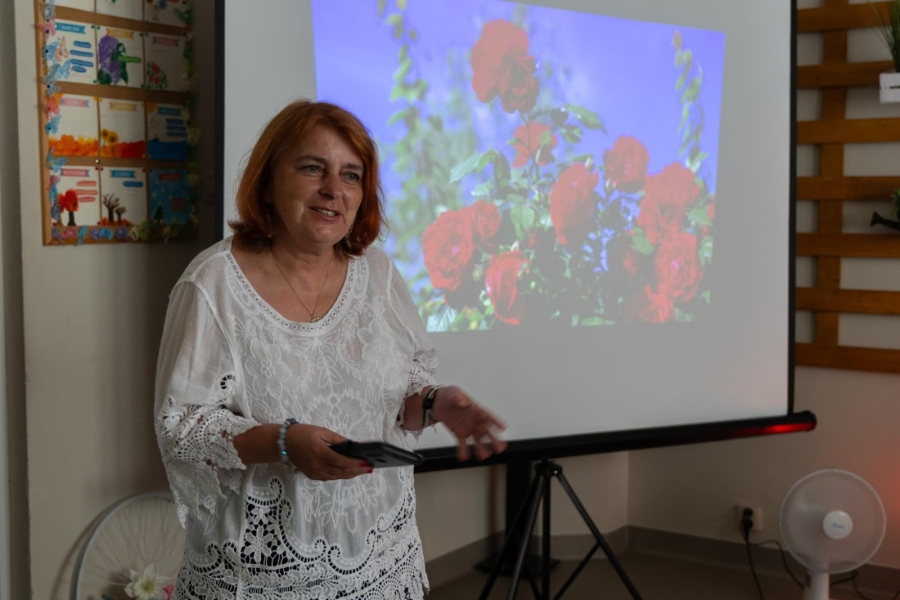 1.Prezes Stowarzyszenia w białej bluzce przemawiająca na tle slajdu z kwiatami