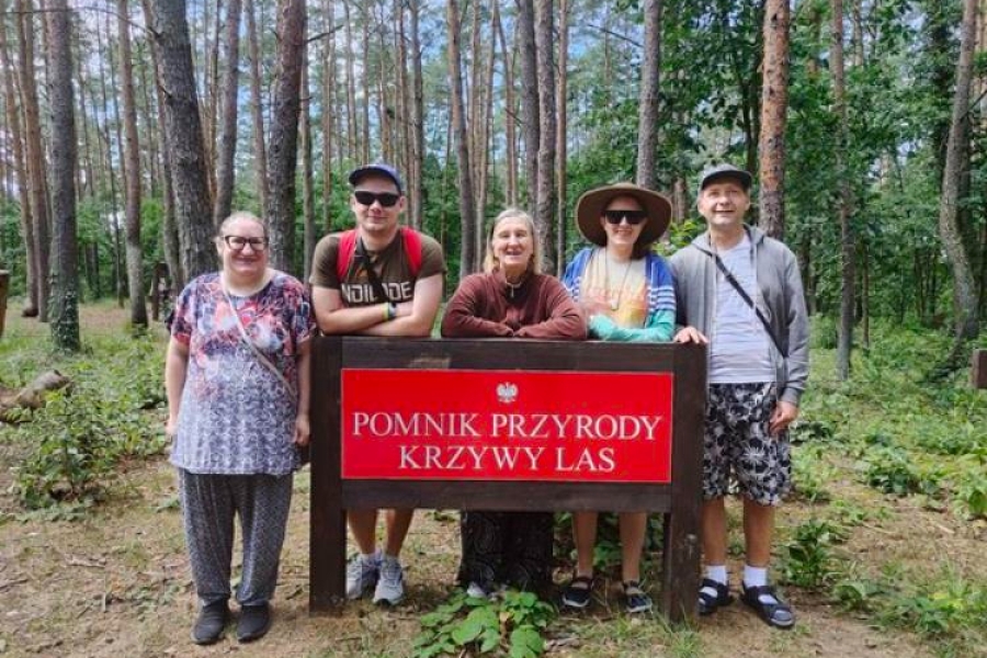 1.Uczestnicy opierający się o tablicę z napisem „Pomnik Przyrody Krzywy Las” na tle drzew