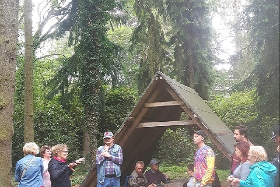 Grupa uczestników odpoczywa w lesie pod drewnianym daszkiem.