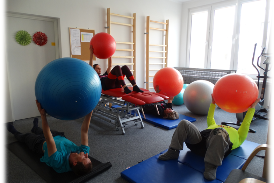 Leżący uczestnicy trzymają w górze piłki na wyprostowanych rękach.