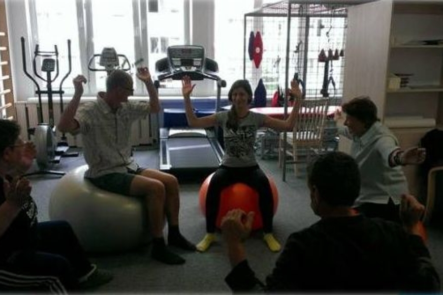 Terapeutka razem z uczestnikami siedzą na piłkach i trzyma zgięte ręce w górze.