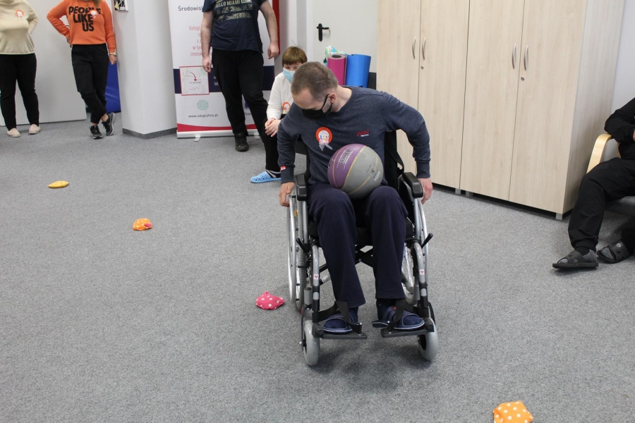 Uczestnik siedzący na wózku inwalidzkim wykonujący slalom tyłem z piłką na kolanach.