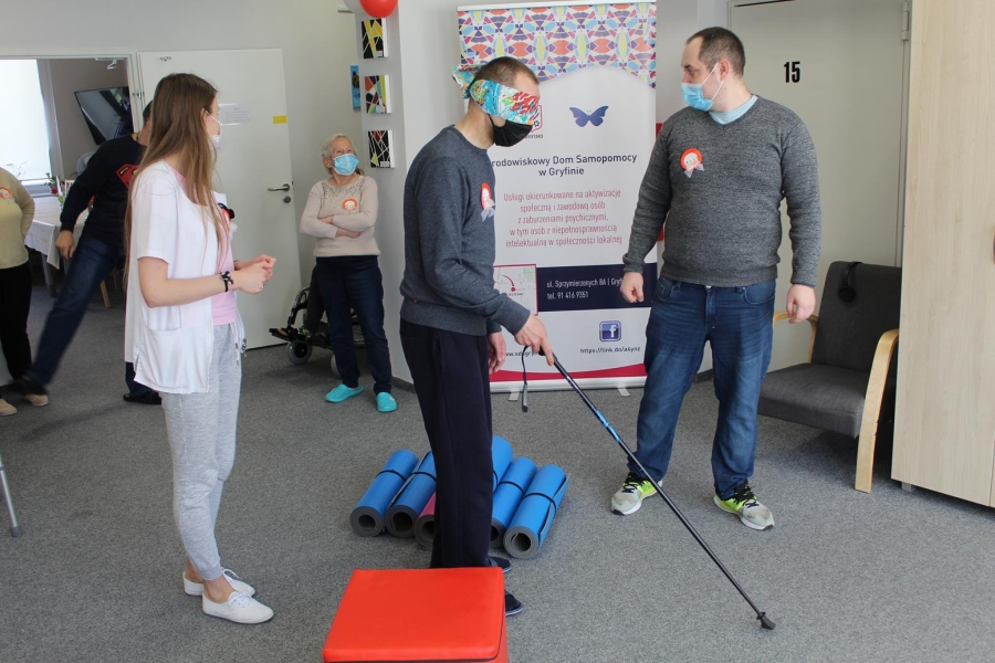 Uczestnik w asyście terapeutów pokonujący tor przeszkód z zasłoniętymi oczami.