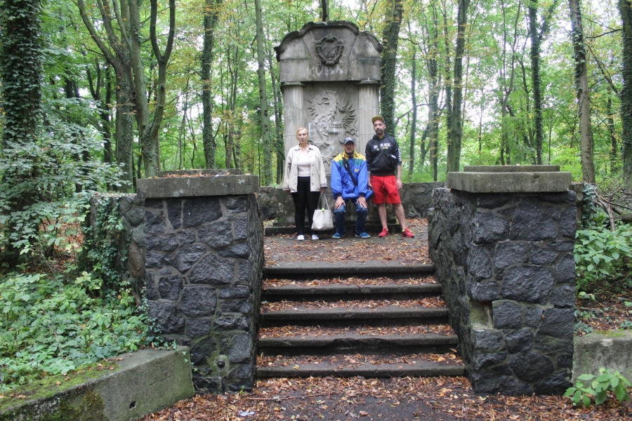 6 – Troje Uczestników pozujących do zdjęcia na tle pomnika.